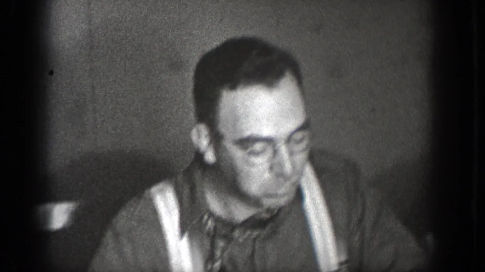 A man in suspenders looks down at the table in black-and-white 1939 home-movie footage from Chelan, Washington.