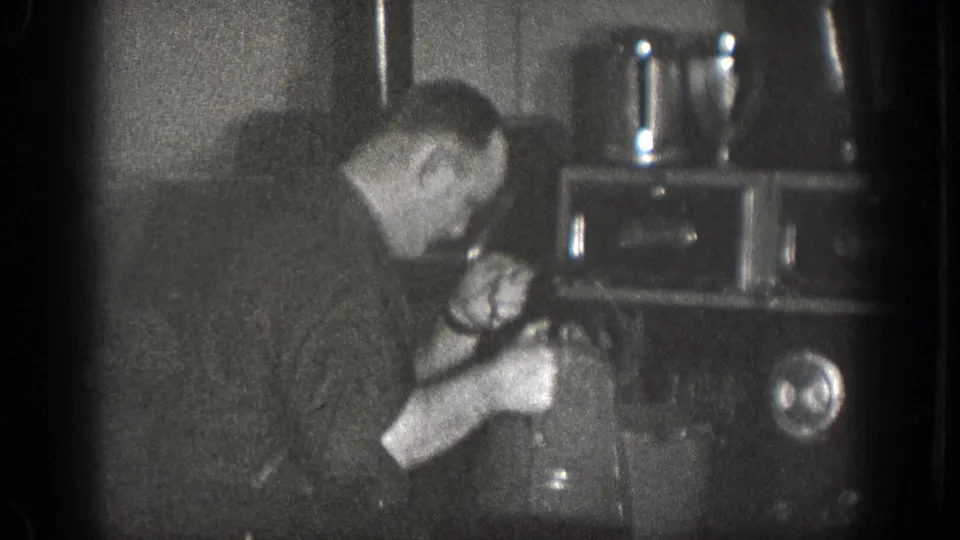 In 1939 Chelan, Washington home-movie footage, a man bends over a stove while working with a kettle and pot in a dark kitchen.