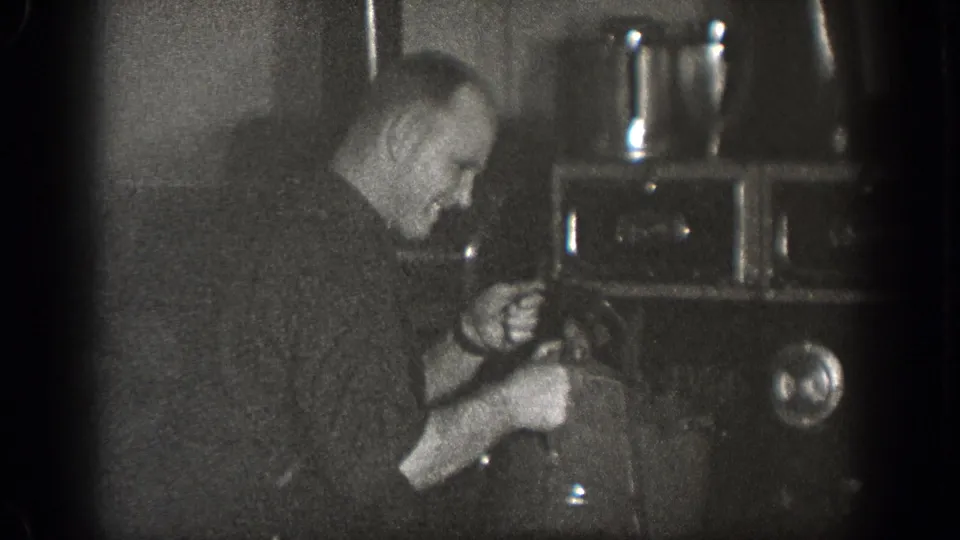 A man in a dark shirt appears to pour or handle liquid from a kettle into a pot on a stove in a 1939 Chelan kitchen scene.