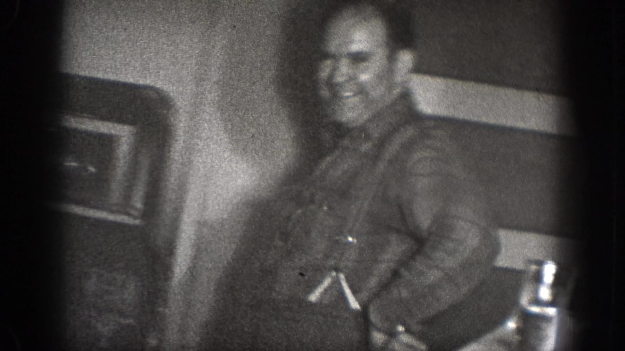A smiling adult man stands near kitchen fixtures in grainy black-and-white 8mm footage from the Chelan, Washington household scene.