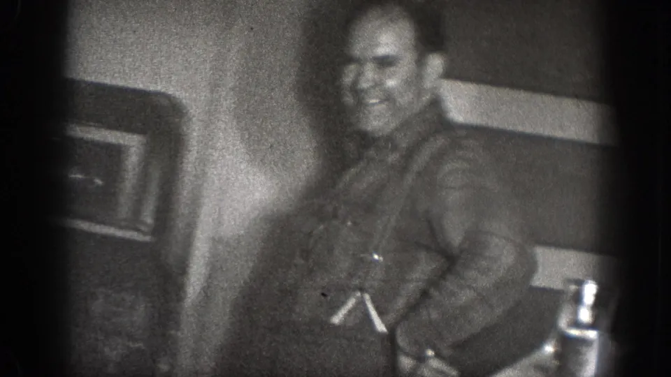 A smiling adult man stands near kitchen fixtures in grainy black-and-white 8mm footage from the Chelan, Washington household scene.