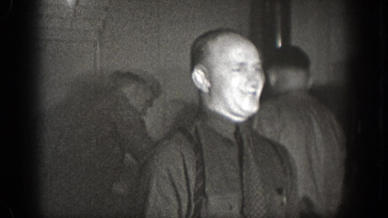 Smiling bald man in a dark shirt and tie standing among other men indoors.