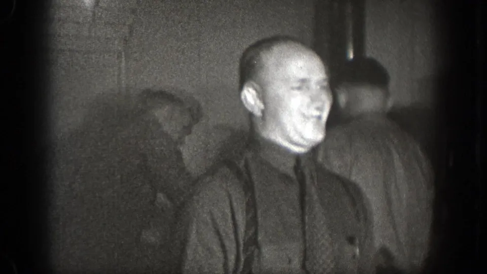 In 1939 Chelan, Washington home-movie footage, a smiling man in a dark shirt and tie stands in a dim interior with other men behind him.