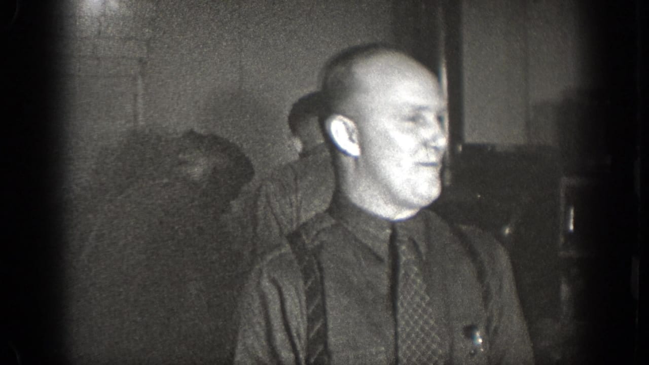 The Chelan, Washington 1939 home-movie frame shows a man in a dark shirt and tie turned in profile, with other men blurred in the background.