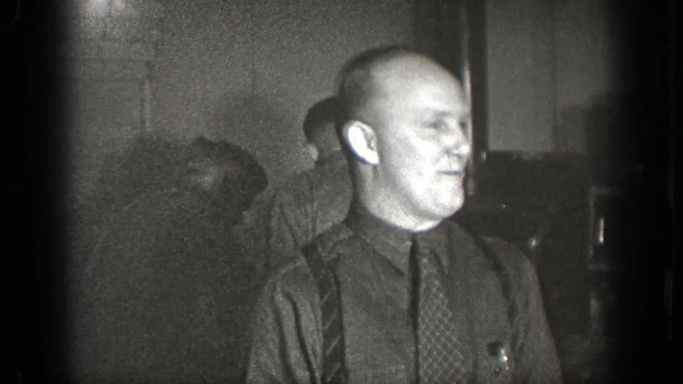 The Chelan, Washington 1939 home-movie frame shows a man in a dark shirt and tie turned in profile, with other men blurred in the background.