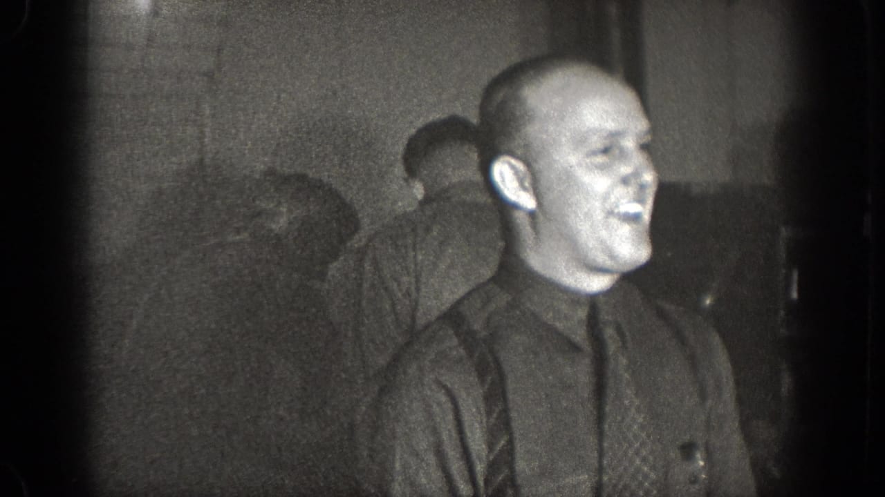 A laughing man in a dark shirt and tie appears in this 1939 Chelan home-movie frame, surrounded by other adult men in a low-lit interior.