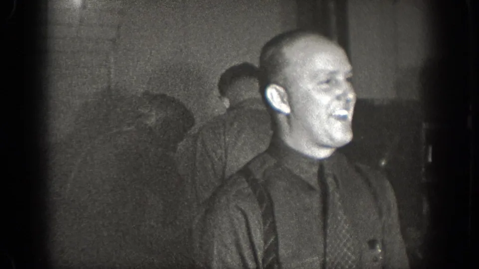A laughing man in a dark shirt and tie appears in this 1939 Chelan home-movie frame, surrounded by other adult men in a low-lit interior.