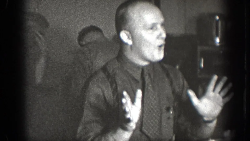 In the final 1939 Chelan frame, a man in a dark shirt and tie raises both hands while speaking or laughing among other men indoors.