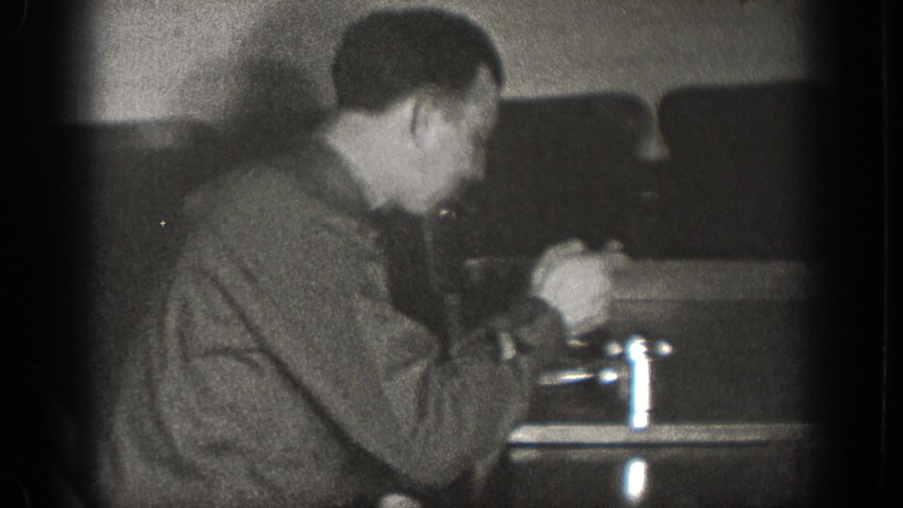 The man leans toward the table while handling food or a utensil, with metal cups and chairs visible in the 1939 interior scene.