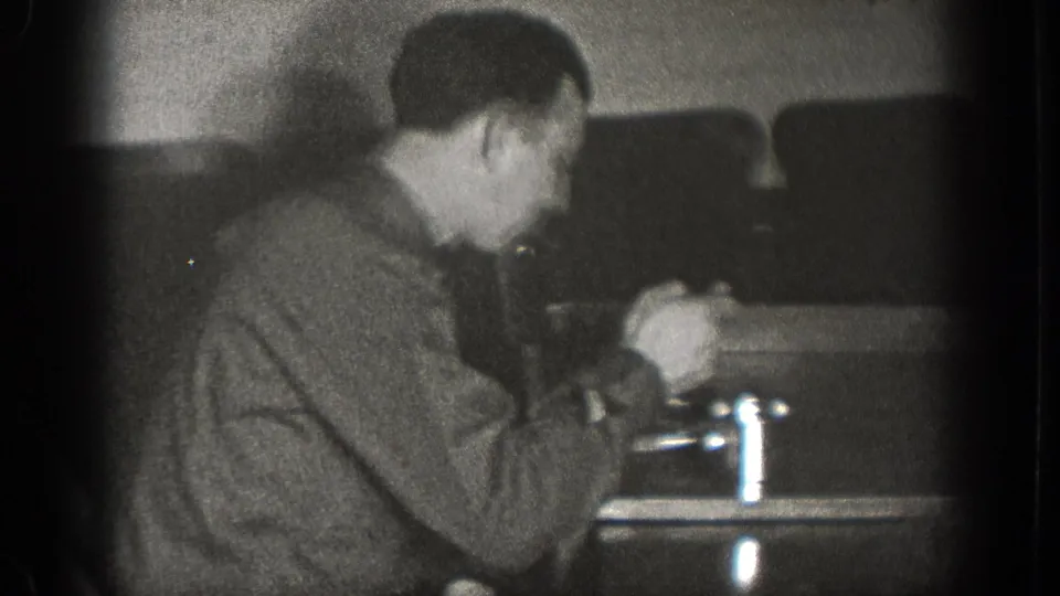 The man leans toward the table while handling food or a utensil, with metal cups and chairs visible in the 1939 interior scene.