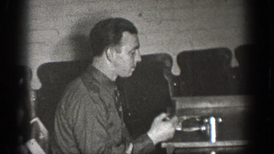 A man sits in profile at a lunch table in the Chelan, Washington home movie, with cups and upholstered seating behind him.