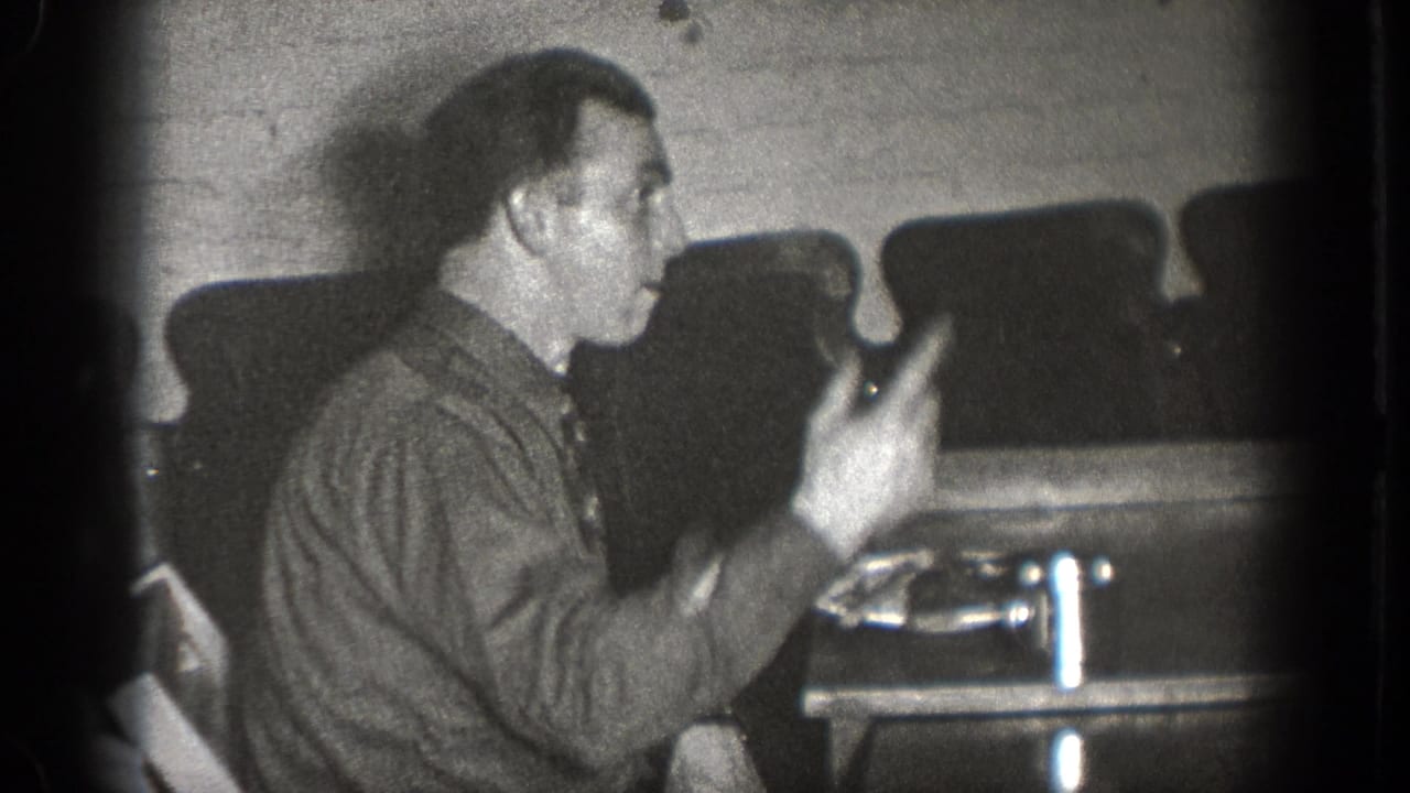 In 1939 Chelan home-movie footage, a seated man in a button-up shirt gestures with a fork near a table in a dim interior.