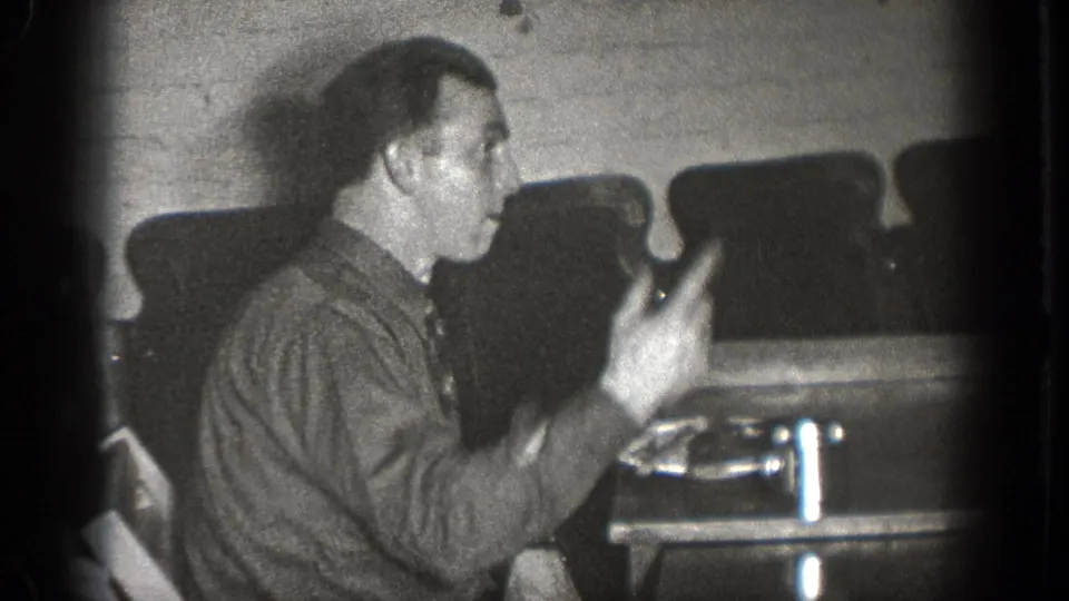 In 1939 Chelan home-movie footage, a seated man in a button-up shirt gestures with a fork near a table in a dim interior.