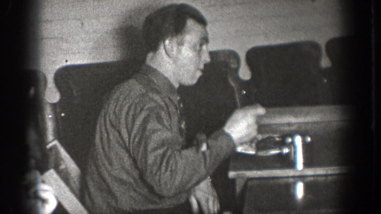 A man at a Chelan, Washington indoor gathering gestures with a fork beside plates and chairs in grainy 1939 8mm footage.