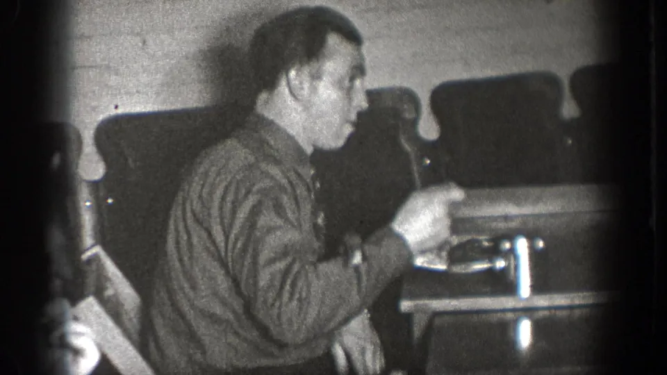 A man at a Chelan, Washington indoor gathering gestures with a fork beside plates and chairs in grainy 1939 8mm footage.