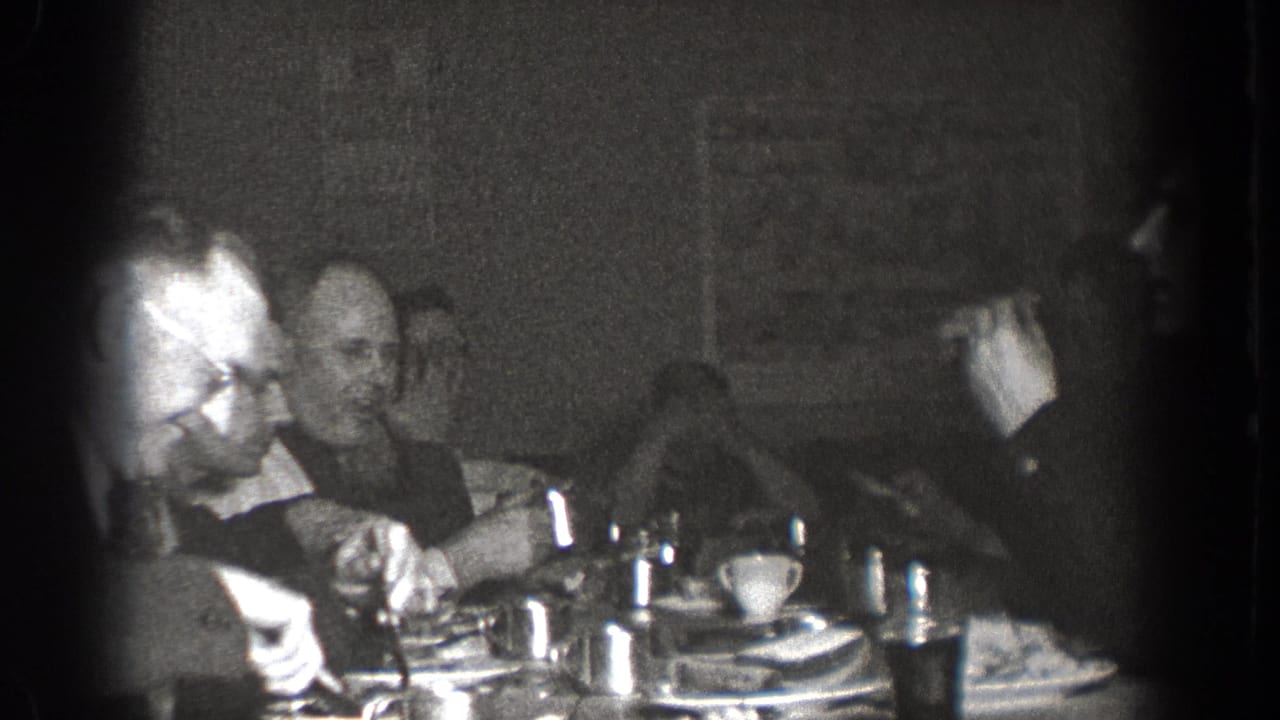 Adults sit around a table with dishes and cups during a 1939 Chelan social gathering, filmed in grainy black-and-white 8mm.