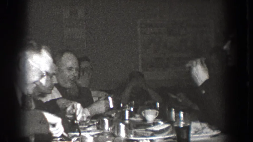 Adults sit around a table with dishes and cups during a 1939 Chelan social gathering, filmed in grainy black-and-white 8mm.