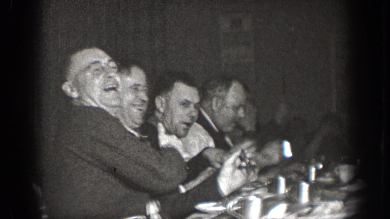In a 1939 Chelan home-movie frame, suited men laugh and gesture together at a table set with dishes and cups.