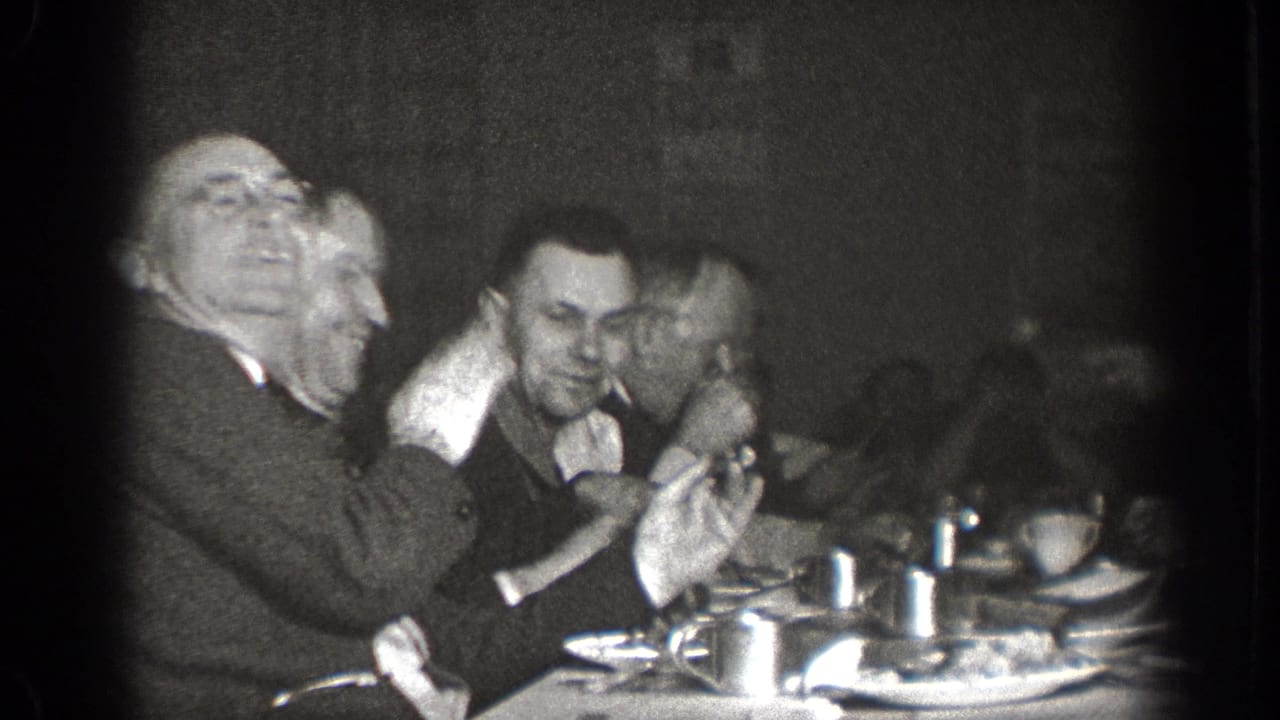 In 1939 Chelan, Washington home-movie footage, adult men sit closely around a table with cups and plates as one man gestures animatedly.