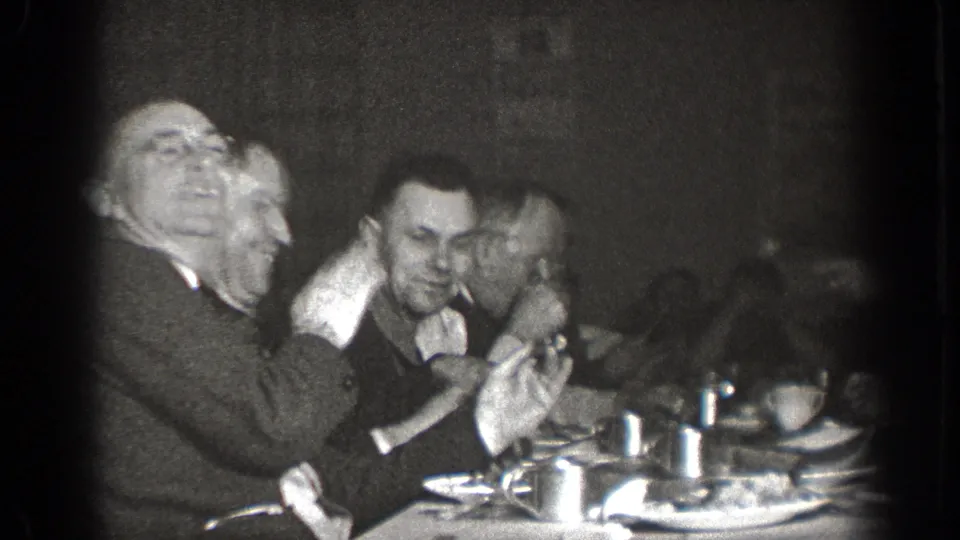 In 1939 Chelan, Washington home-movie footage, adult men sit closely around a table with cups and plates as one man gestures animatedly.