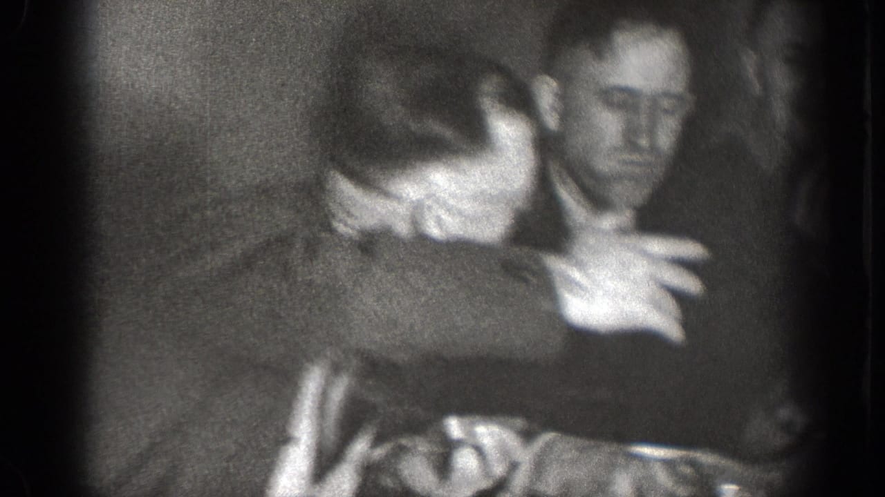 Soft-focus 8mm footage from the Chelan gathering shows adult men leaning together and gesturing during a lively indoor conversation.