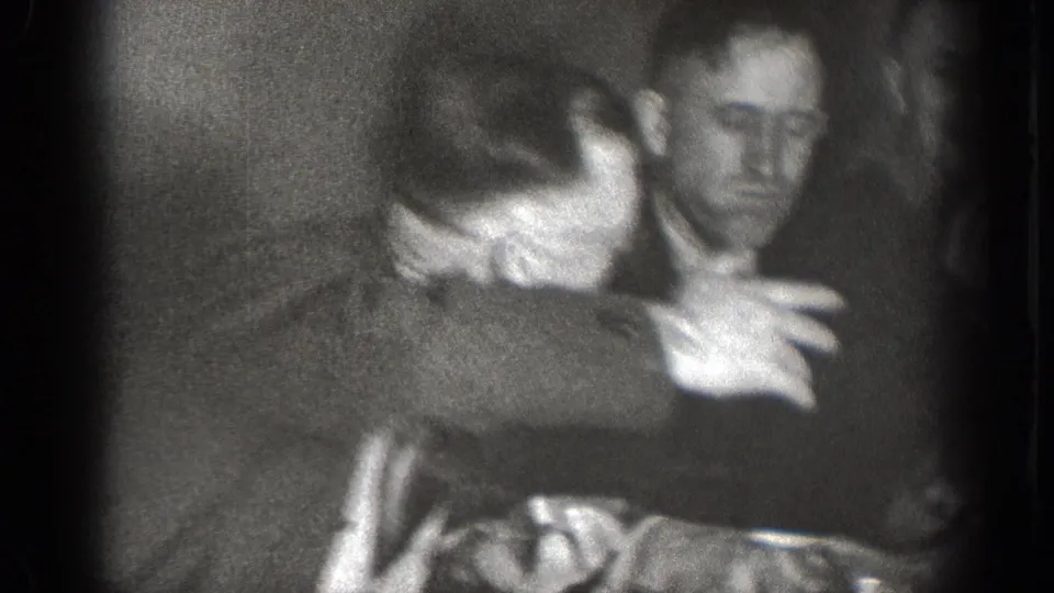 Soft-focus 8mm footage from the Chelan gathering shows adult men leaning together and gesturing during a lively indoor conversation.