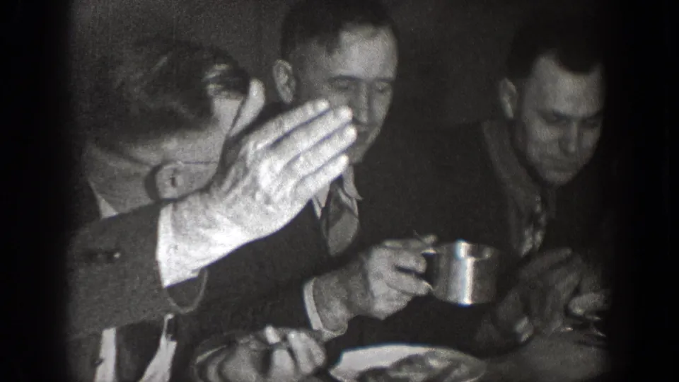 In 1939 Chelan home-movie footage, adult men in suits sit at a dining table with plates and cups in a dim interior.