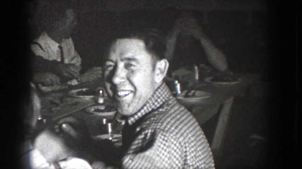A man smiles toward the camera during the Chelan, Washington indoor gathering, with other diners and tableware around him.