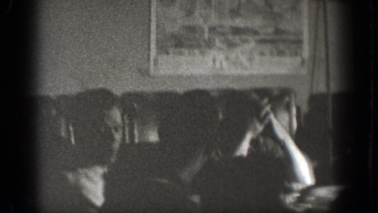 A grainy 1939 Chelan interior frame shows seated figures at a table, with a framed picture visible on the wall behind them.