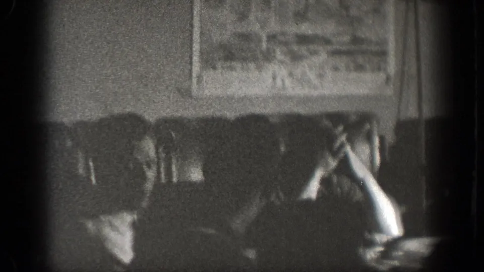 A grainy 1939 Chelan interior frame shows seated figures at a table, with a framed picture visible on the wall behind them.