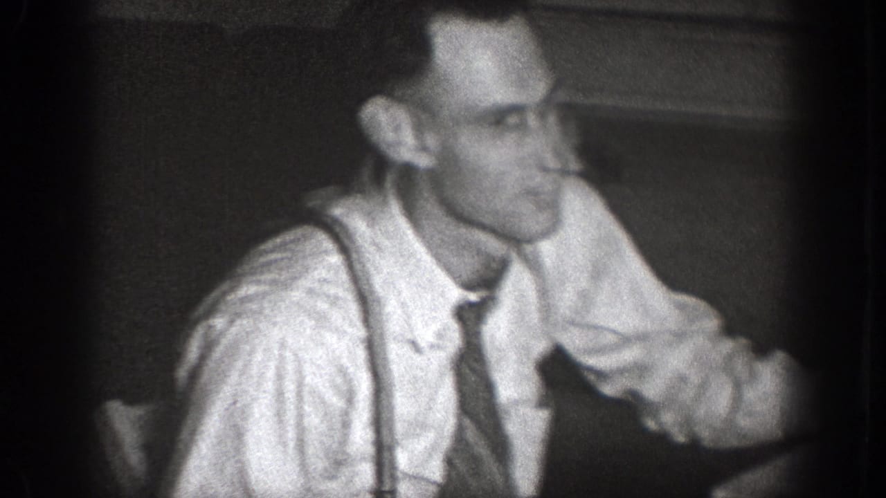Adult man in white shirt, dark tie, and suspenders seated indoors in 1939 Chelan home-movie footage.