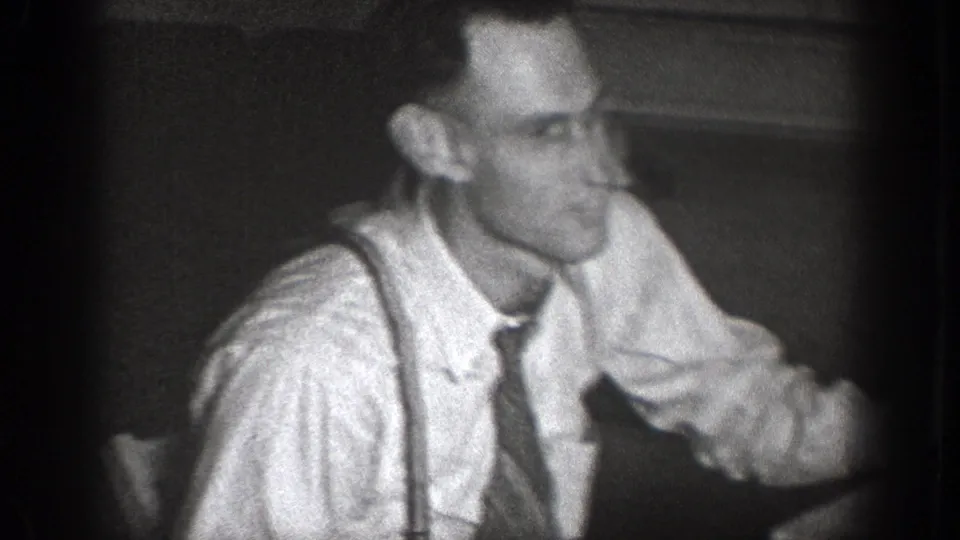 Adult man in white shirt, dark tie, and suspenders seated indoors in 1939 Chelan home-movie footage.