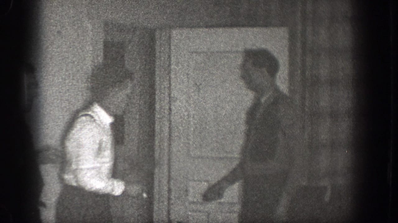 Two adult men stand by an interior door, with the shirt-and-suspenders man facing another figure.