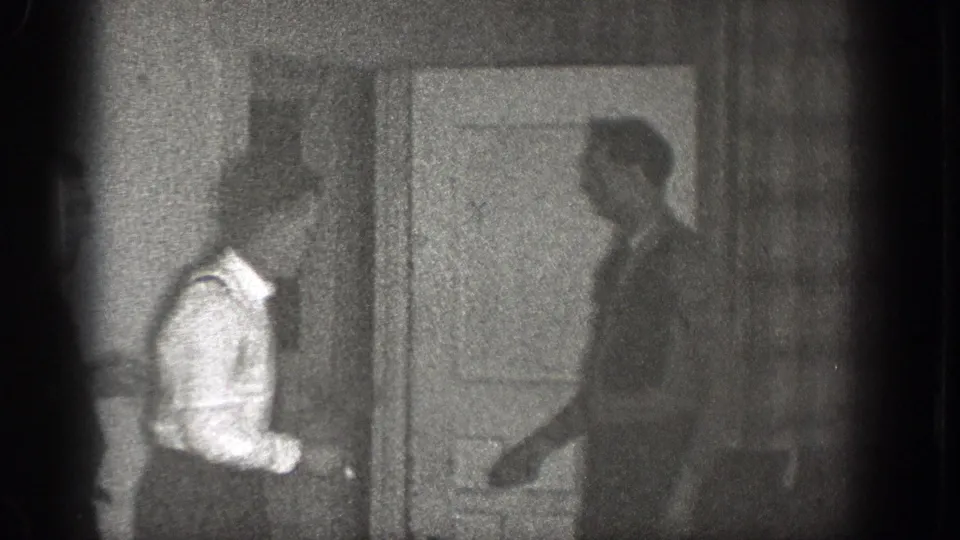 Two adult men stand by an interior door, with the shirt-and-suspenders man facing another figure.