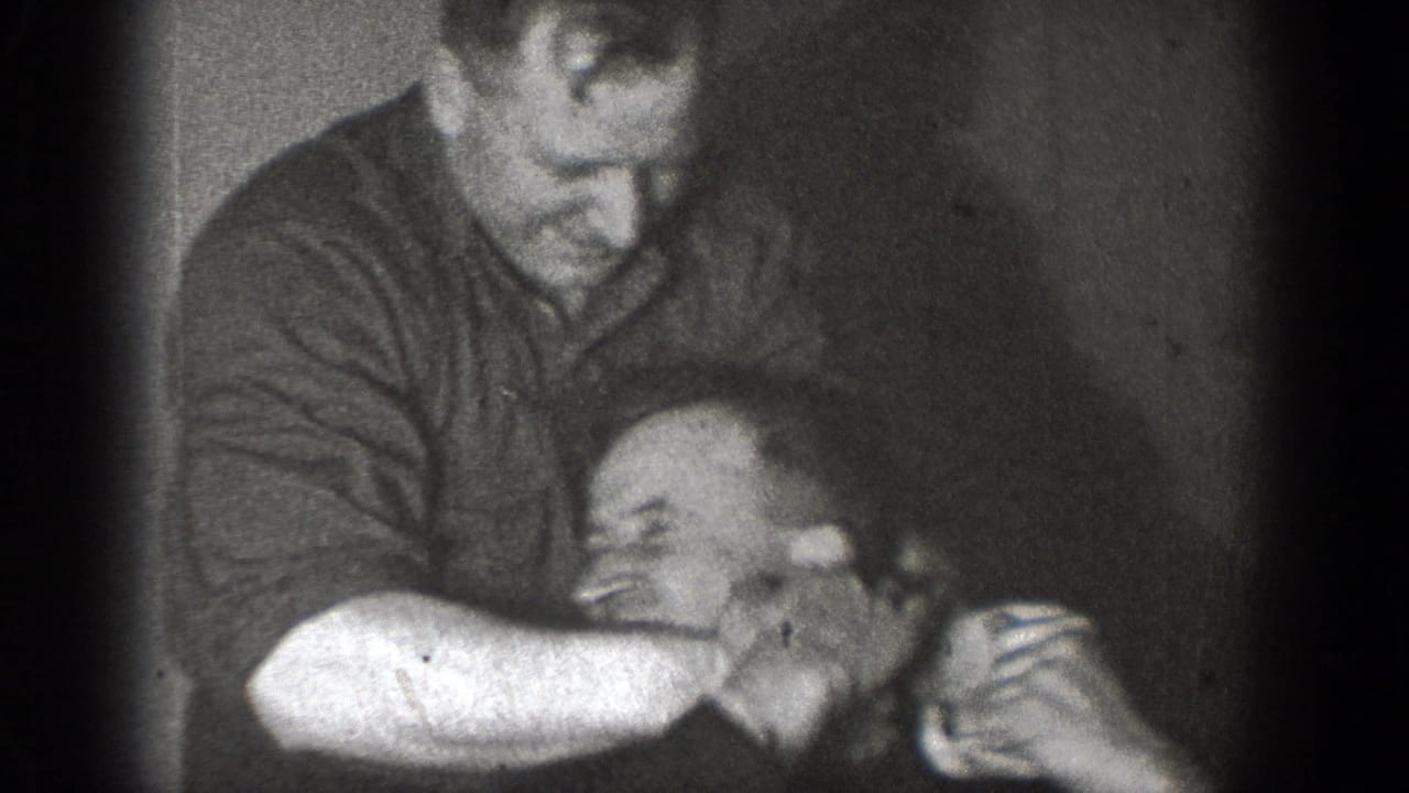 A black-and-white 8mm frame shows men in Chelan, Washington, playfully grappling in a private indoor setting.