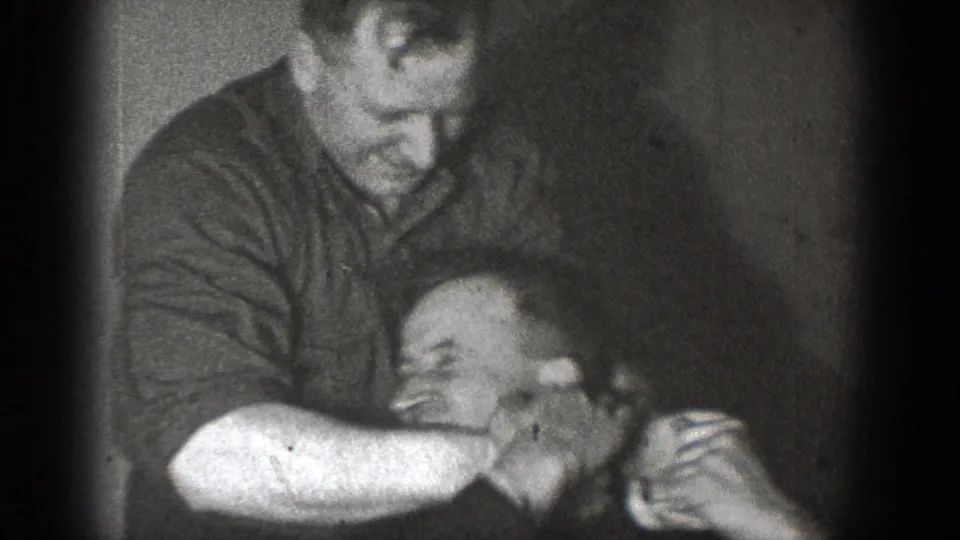 A black-and-white 8mm frame shows men in Chelan, Washington, playfully grappling in a private indoor setting.