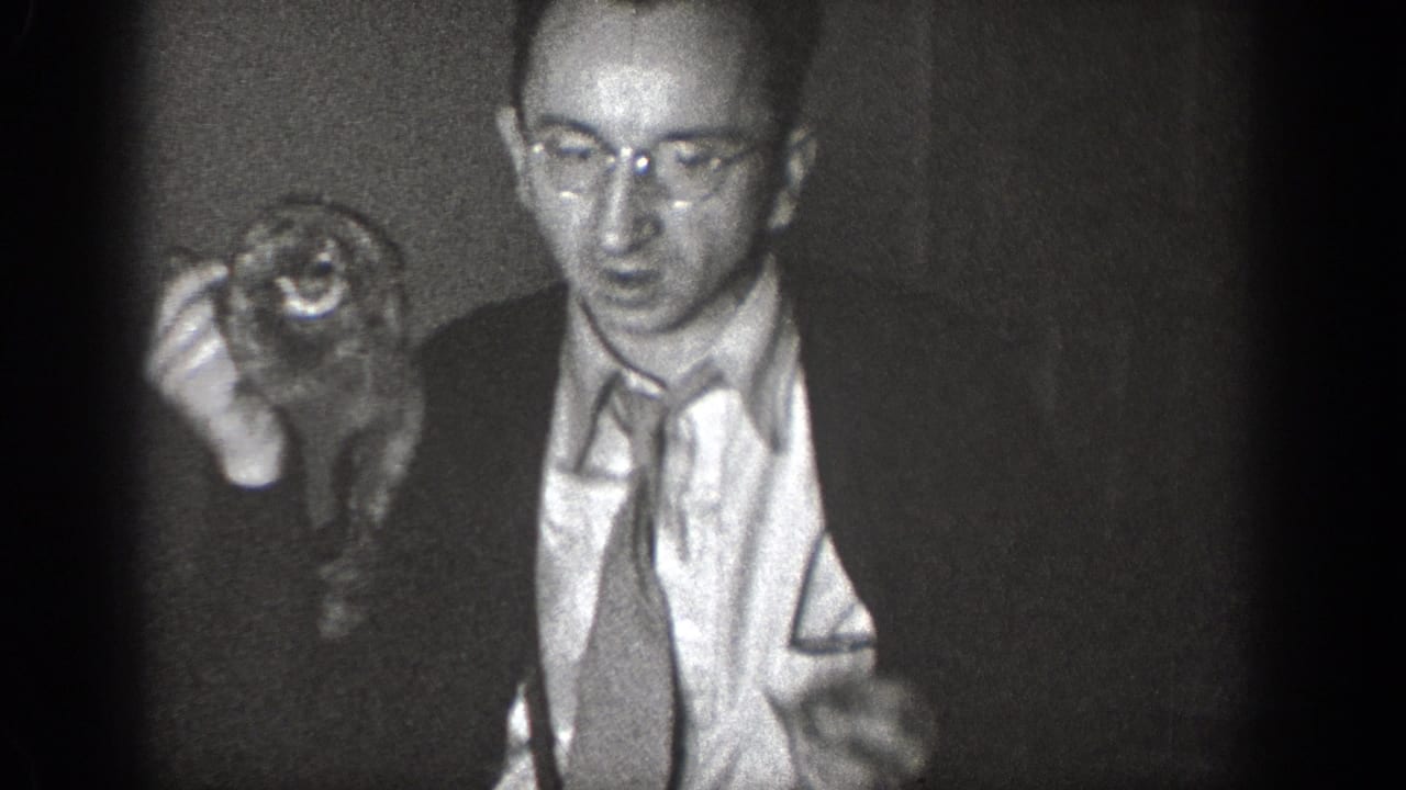 A suited man with glasses holds a small animal close to the camera in grainy 1939 Chelan home-movie footage.