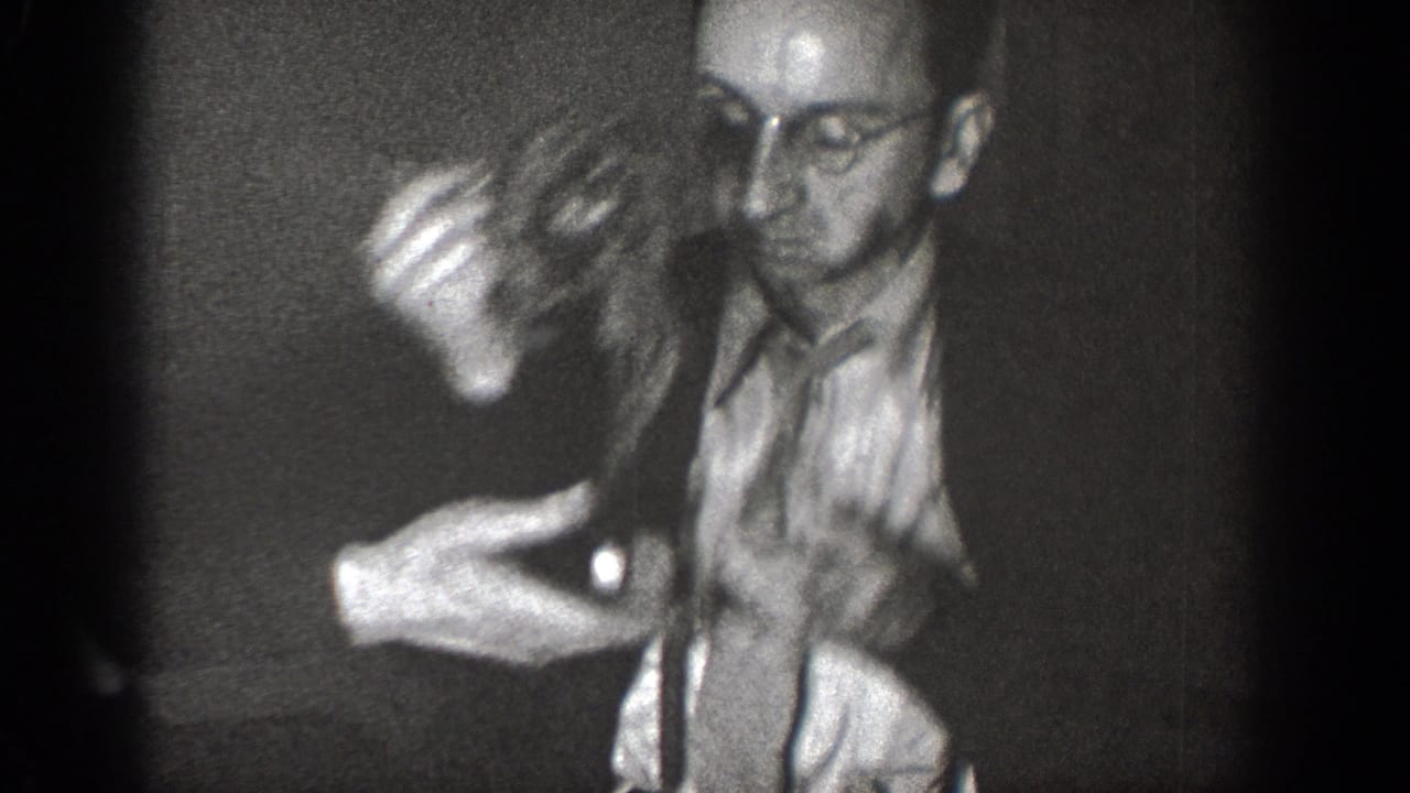 A man in glasses handles a small animal in a dark interior, with another partially visible figure below the frame.