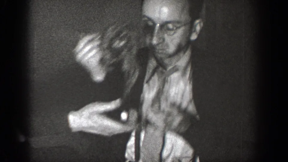 A man in glasses handles a small animal in a dark interior, with another partially visible figure below the frame.