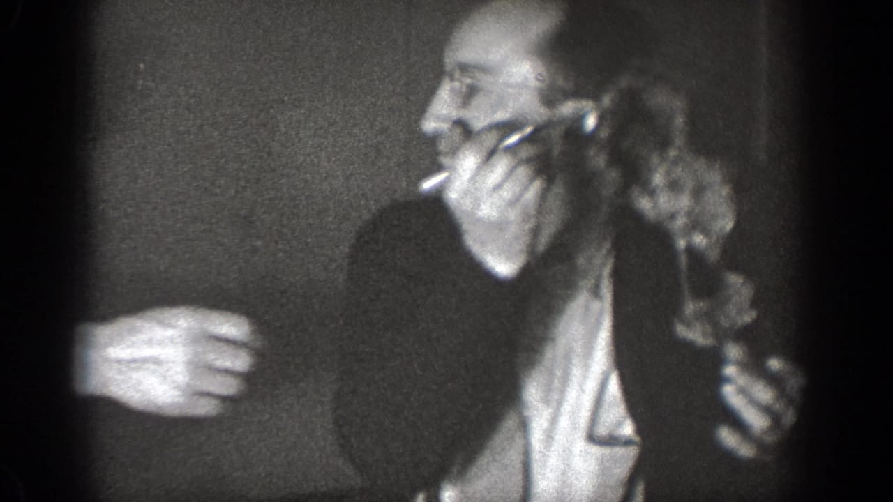 Adult men in suits crowd together in a dim room, one man holding an object near his mouth.