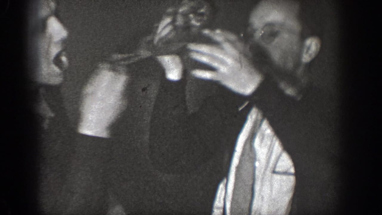 A man holds another man close while a third figure appears between them in shadow.