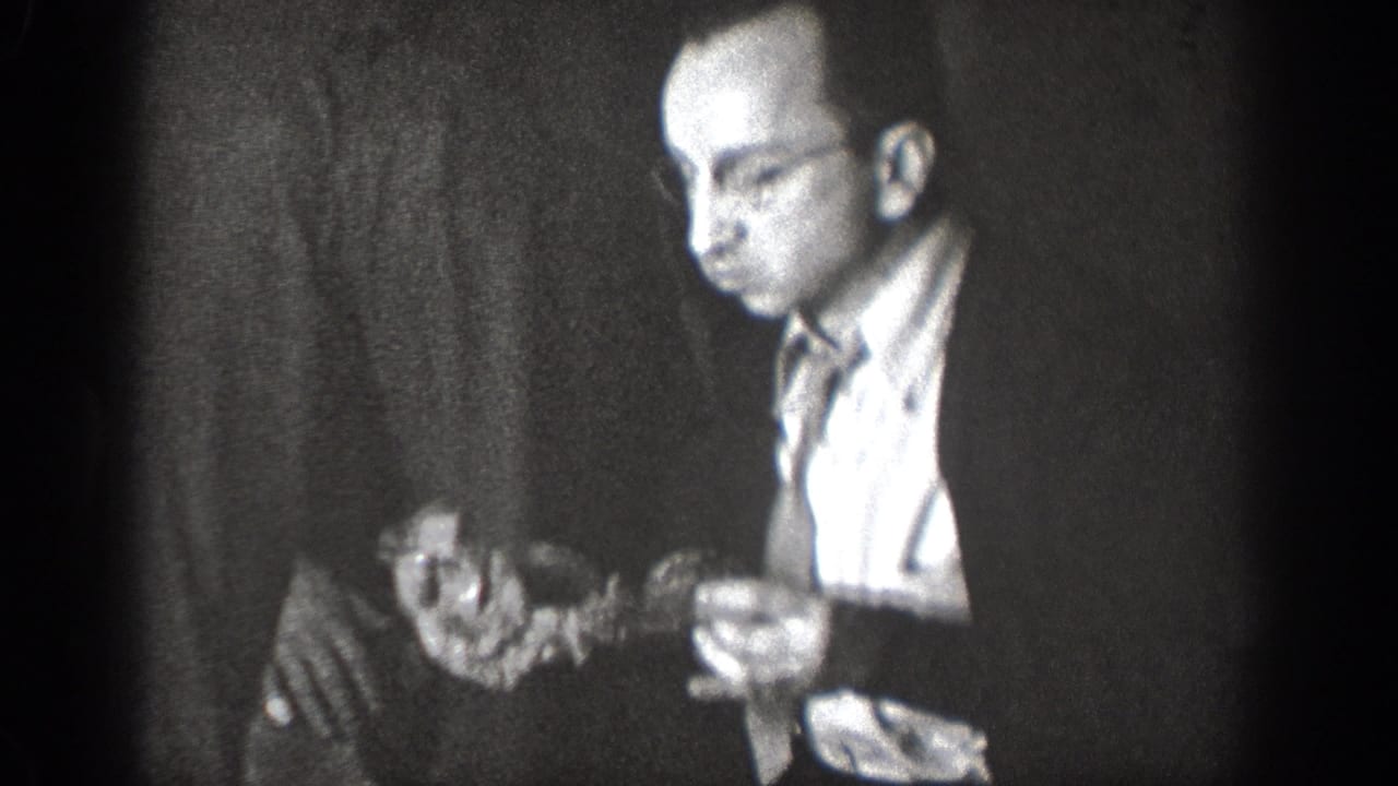In 1939 Chelan home-movie footage, a man in glasses and a dark jacket holds food in a dim indoor gathering.