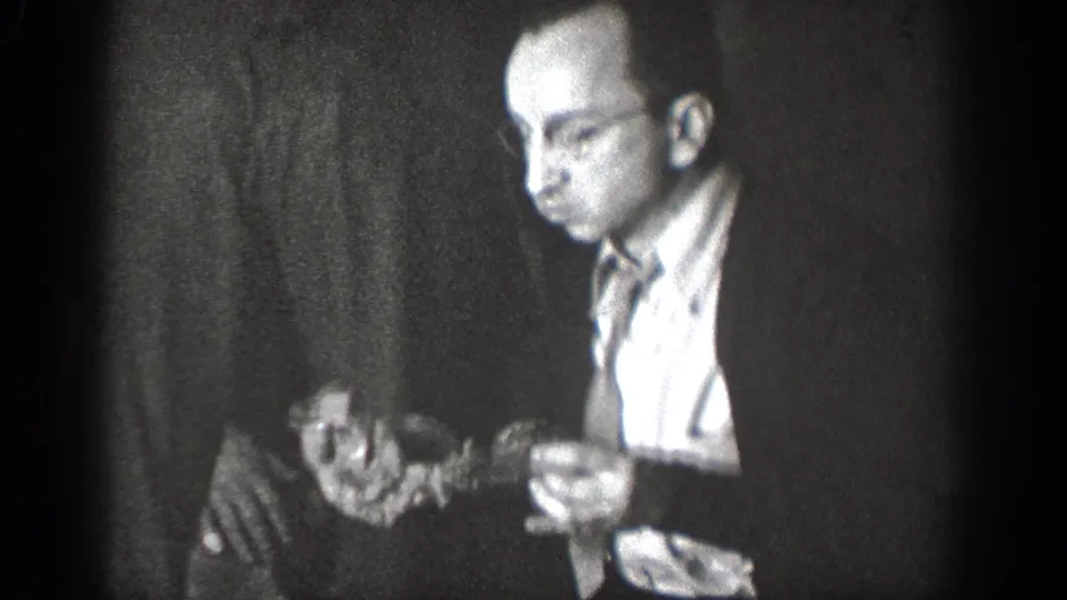 In 1939 Chelan home-movie footage, a man in glasses and a dark jacket holds food in a dim indoor gathering.