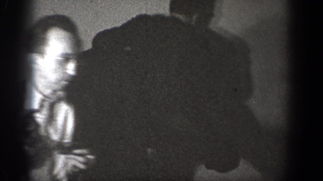Blurred man in glasses stands near shadowed figures in a crowded room.