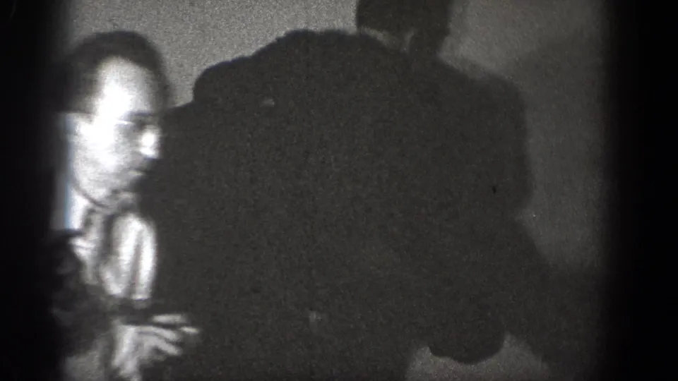 A blurred frame from the Chelan 1939 home movie shows a man in glasses among shadowed men in a private interior.