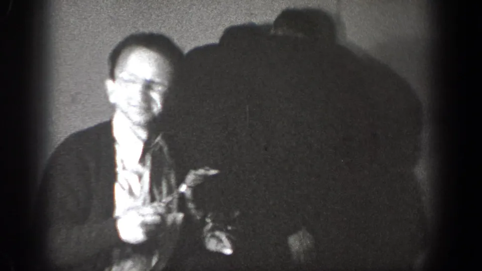 The man in glasses faces the camera in 1939 Chelan footage, holding food as another figure leans in nearby.