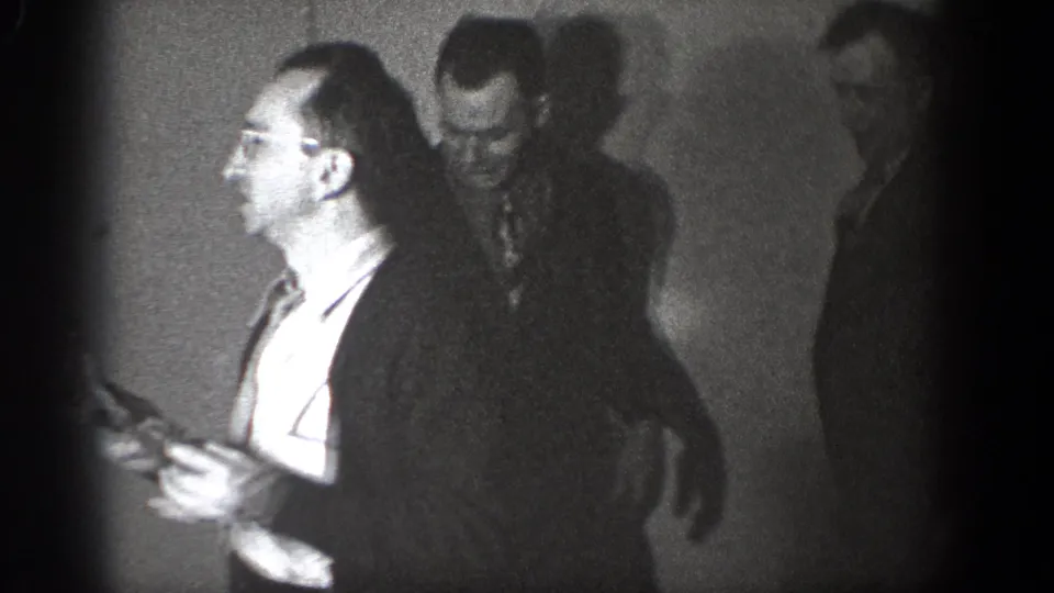 In the final frame, the man in glasses appears in profile while two men stand behind him in the Chelan indoor gathering.