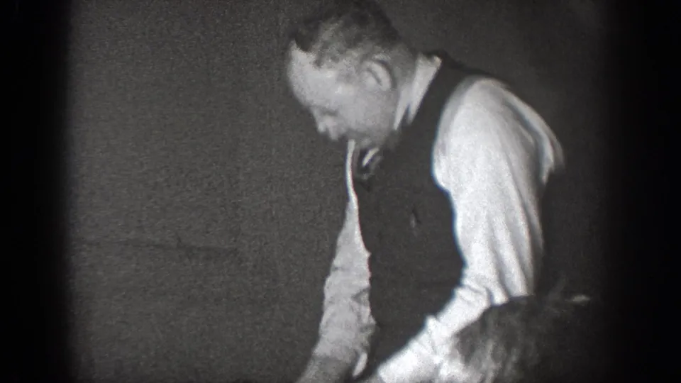 In a 1939 Chelan, Washington home-movie frame, a man in a vest and dress shirt appears to work over a table indoors.