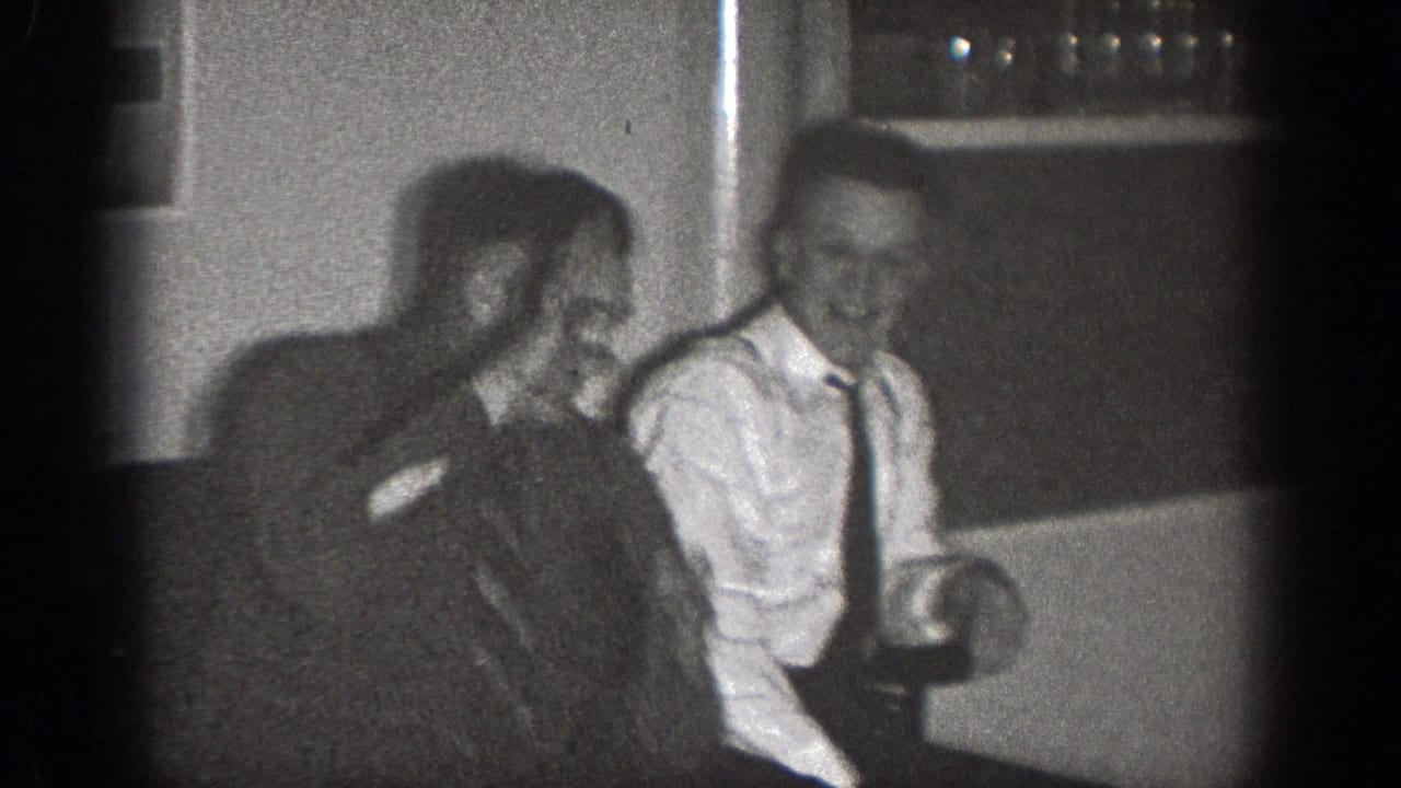 Two adult men sit together indoors, one in a dark jacket and one in a light shirt and tie.
