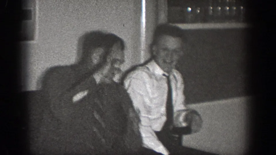 Two adult men sit close together indoors in Chelan, Washington footage from 1939, one laughing beside a man in a shirt and tie.